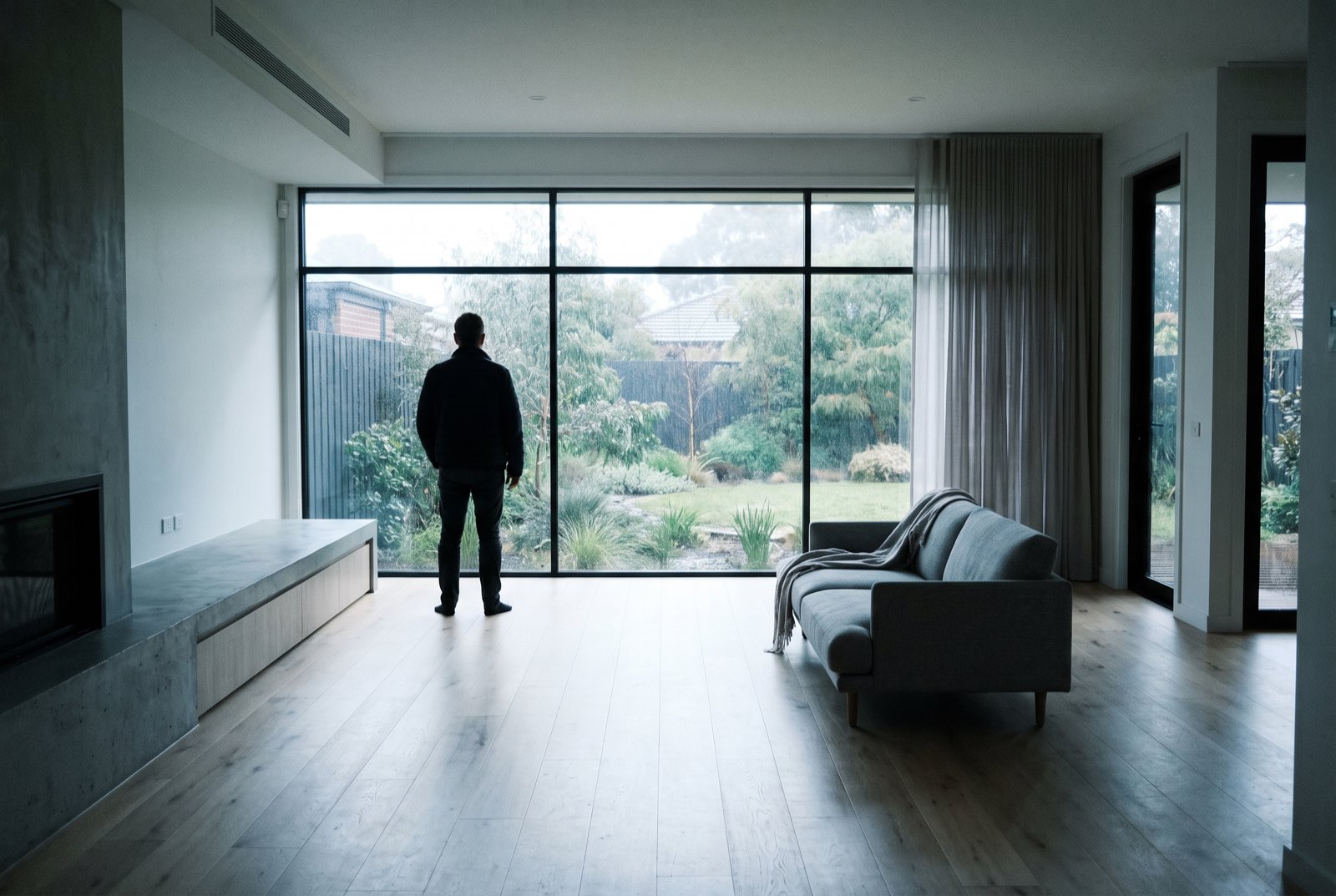 A single person standing alone at the window of an empty open home inspection in a modern Australian living room